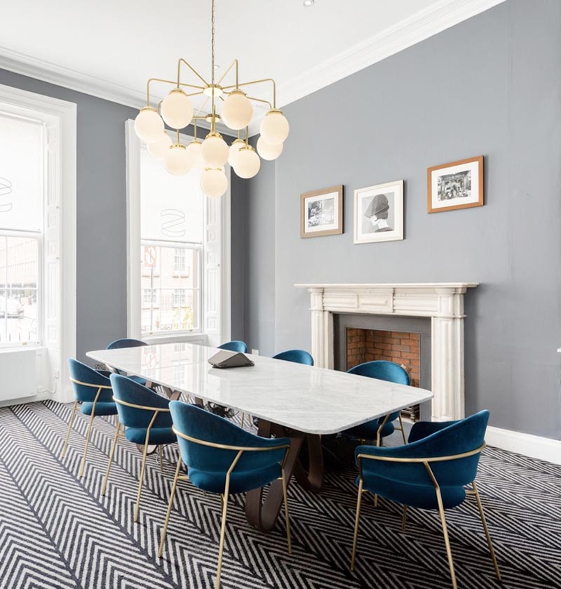 Modern dining room with a marble table, blue velvet chairs, a geometric chandelier, a fireplace, and three framed photos on a grey wall with chevron-patterned carpet. contact insight