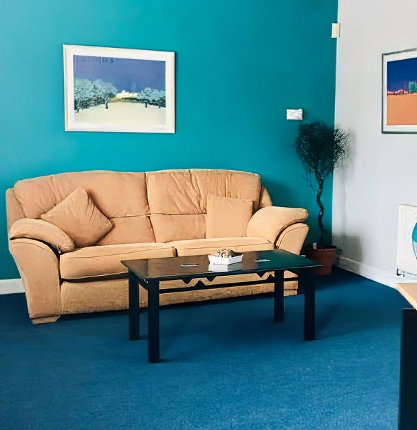 A tan sofa with two cushions sits against a teal wall, framed art above offering contact insight, and a black coffee table in front on blue carpet. A potted plant is in the corner.