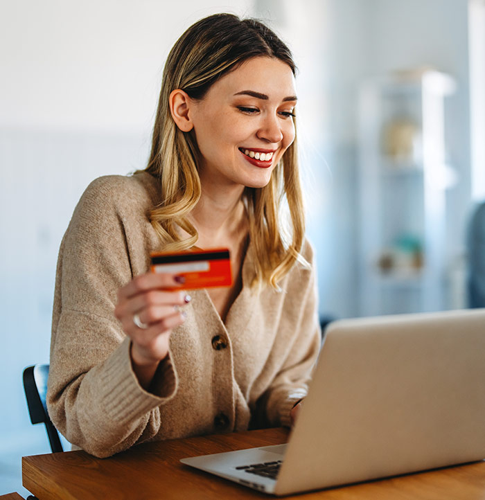 A woman sits at a table holding a credit card and using a laptop, appearing to make an online purchase on popular e-commerce websites.