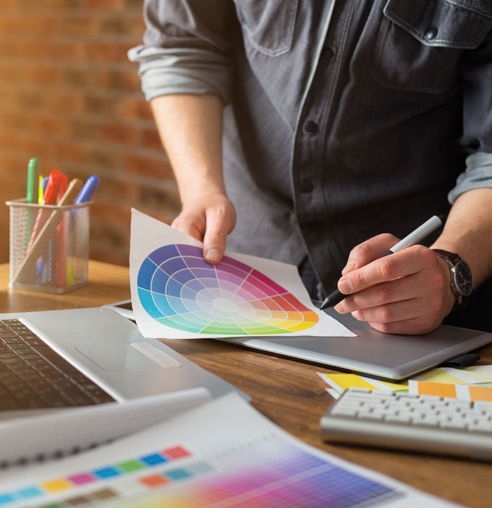 Person reviewing a colour wheel chart while using a pen tablet at a desk with a laptop, colour swatches, and stationery during the website redesign process.